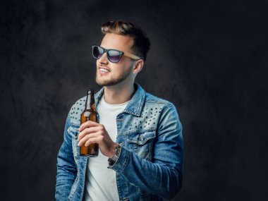 Pozitif sarışın erkek denim ceket ve güneş gözlüğü giymiş bir zanaat bira şişesiyle tutar.