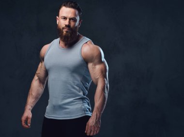 Gri arka plan üzerinde izole bir kolsuz gömlek giymiş acımasız sakallı vücut geliştirmeci portresi.