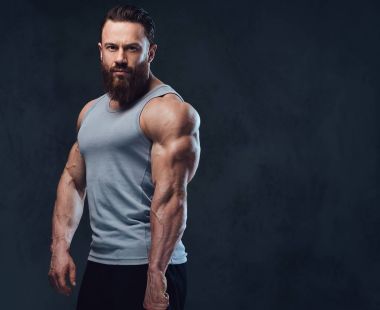 Gri arka plan üzerinde izole bir kolsuz gömlek giymiş acımasız sakallı vücut geliştirmeci portresi.