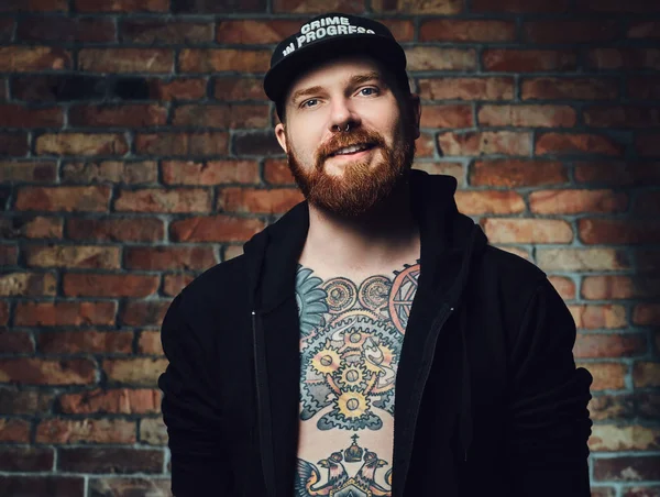 Kızıl saçlı sakallı erkek silah ve göğsünde, dövmeli portresi giymiş siyah spor ceketli bir tuğla duvarın üzerinden.