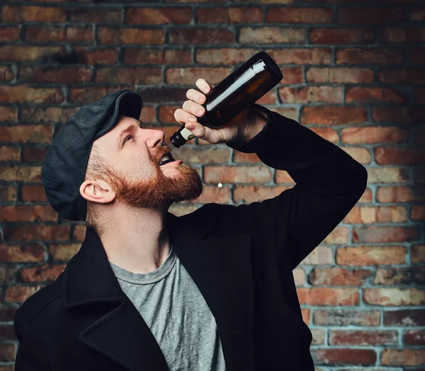 Bearded male dressed in a black tweed flat cap and wool jacket holds beer bottle over the wall of a brick.