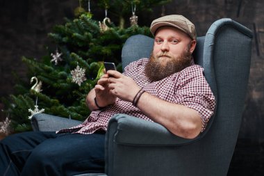 Bir sandalye ve bir odada Noel dekorasyonu ve hediyeler ile smartphone kullanarak tombul sakallı hippi erkek oturur.