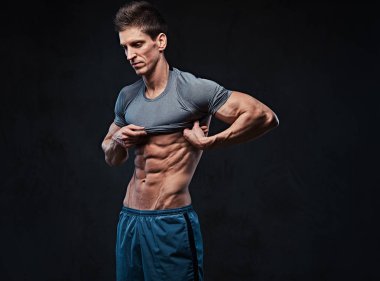 Stüdyo portre ectomorph kas gömleksiz erkek t gömlek alır ve göğüs kasları koyu gri arka plan üzerinde gösterir.