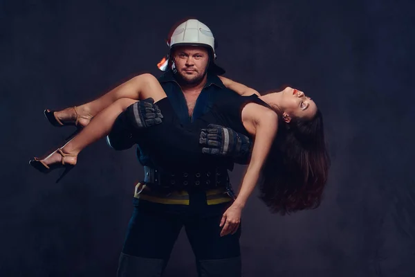 Brutal firefighter holds saved brunette female dressed in a black evening dress.