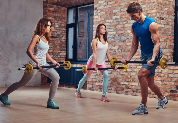 Two fitness women and a coach, stretching squats with barbells in a club with loft interior.