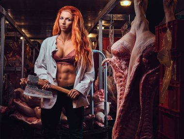 The redhead athletic female holds an ax in a meat factory.
