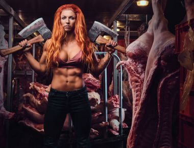The redhead athletic female holds two axes in a meat factory.