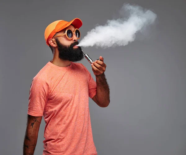 在橙色的帽子和太阳镜吸烟的一个长胡子的时髦男性的肖像抽烟电子管道 灰色背景下的隔离 — 图库照片