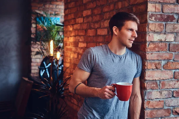 ジーンズや シャツに身を包んだスタイリッシュな Tattoed 魅力的な男は一杯のコーヒーを保持しているレンガの壁に対して傾きます 笑みを浮かべて よそ見 — ストック写真