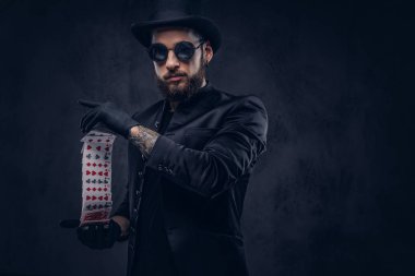 Magician in a black suit, sunglasses and top hat, showing trick with playing cards on a dark background. clipart