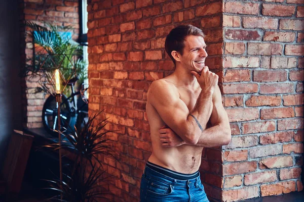 A sexy tattoed shirtless man leans against the brick wall. Smiling and looking away.
