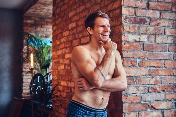 A sexy tattoed shirtless man leans against the brick wall. Smiling and looking away.