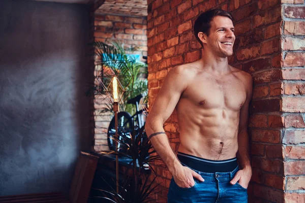 A sexy tattoed shirtless man leans against the brick wall. Smiling and looking away.