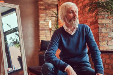 Kazak ve kot gri sakallı yakışıklı bir yaşlı adam çatı iç bir tuğla duvara bir odada oturur.