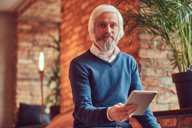 Kazak ve tablet kullanma kot gri sakallı yakışıklı bir yaşlı adam çatı iç bir tuğla duvara bir odada oturur.