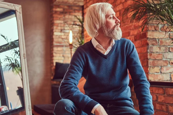 A handsome old man with a gray beard in sweater and jeans sits in a room with loft interior against a brick wall.