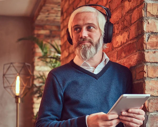 Portrait of an old man with a gray beard in sweater using a tablet and headphones leaning against a brick wall in a room with loft interior.