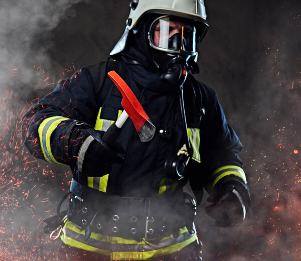 A firefighter dressed in a uniform and an oxygen mask holds a red axe standing in fire sparks and smoke over a dark background.