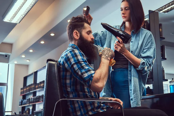 Handsome stylish bearded male with a tattoo on arm dressed in a flannel shirt drinks juice while getting a haircut.