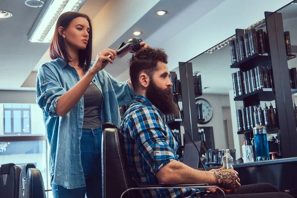 Handsome stylish bearded male with a tattoo on arm dressed in a flannel shirt holding juice while barber female uses a hair-dryer in a barbershop.