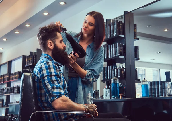Handsome stylish bearded male with a tattoo on arm dressed in a flannel shirt holding juice while barber female uses a hair-dryer in a barbershop.