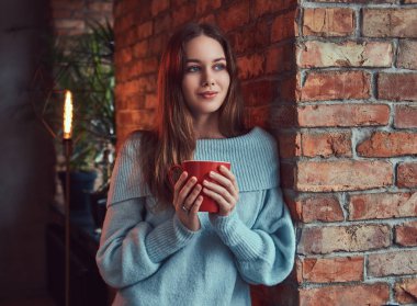 Şehvetli bir esmer gri süveterli çatı iç ile bir odada duvara yaslanmış kahve içer.