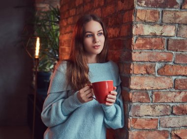 Şehvetli bir esmer gri süveterli çatı iç ile bir odada duvara yaslanmış kahve içer.