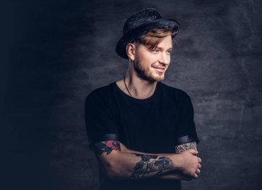 Çapraz dövme kollu yakışıklı sakallı hippi portresi bir siyah t-shirt ve şapka giymiş. Karanlık bir arka plan üzerinde izole.