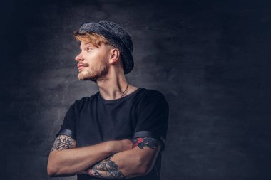 Çapraz dövme kollu yakışıklı sakallı hippi portresi bir siyah t-shirt ve şapka giymiş. Karanlık bir arka plan üzerinde izole.