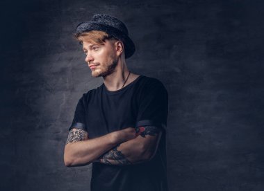 Çapraz dövme kollu yakışıklı sakallı hippi portresi bir siyah t-shirt ve şapka giymiş. Karanlık bir arka plan üzerinde izole.