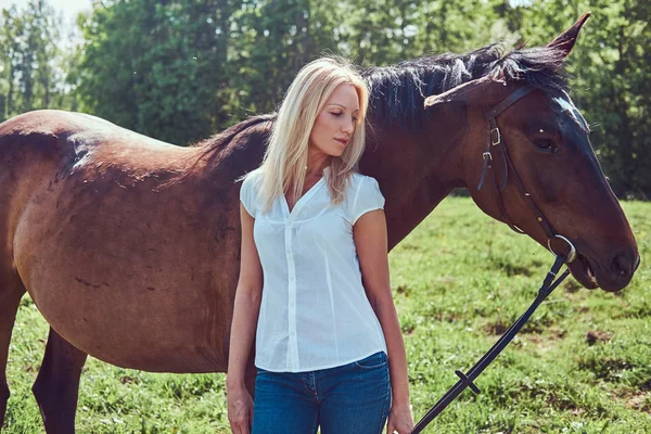 白いブラウスと田舎で馬と立っているジーンズを着て魅力的な美しいブロンド — ストック写真