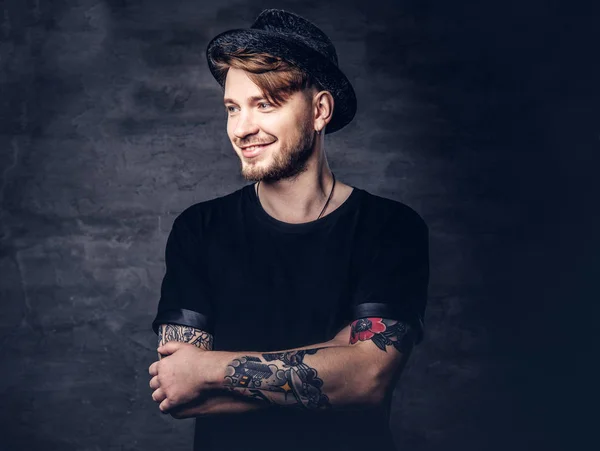 Portrait of handsome bearded hipster with crossed tattooed arms, dressed in a black t-shirt and hat. Isolated on a dark background.