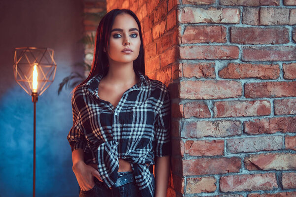 Portrait of an attractive hipster girl wearing flannel shirt leaning on the wall in a room with loft interior.