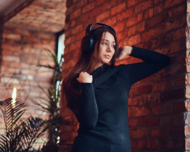 Yakın çekim portre Siyah kazaklı çatı iç ile bir odada bir tuğla duvara yaslanmış iken iyi bir kulaklık ile müzik dinlemek güzel bir sevimli esmer.