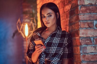 Yakın çekim portre çekici bir esmer çatı iç ile bir odada duvara yaslanmış bir telefon tutarak pazen gömlek giymiş.