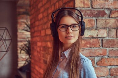 Bir çekici çekici esmer çatı tasarımı ile bir odada bir tuğla duvara karşı yaslanmış kulaklıklar müzik dinlemek gözlük ve mavi gömlekli portresi.