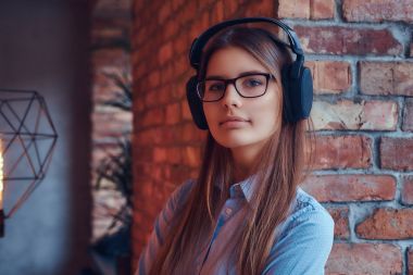 Bir çekici çekici esmer çatı tasarımı ile bir odada bir tuğla duvara karşı yaslanmış kulaklıklar müzik dinlemek gözlük ve mavi gömlekli portresi.