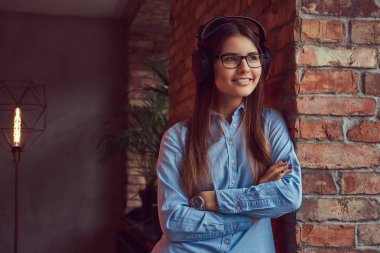 Bir çekici esmer gözlük ve çarpı işareti mavi gömlek kol büyüleyici ve çatı tasarımı ile bir odada bir tuğla duvara karşı yaslanmış kulaklıklar müzik dinlemek portre.