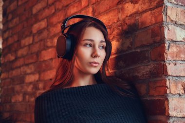 Yakın çekim portre Siyah kazaklı bir güzel şirin esmer çatı iç ile bir odada bir tuğla duvara yaslanmış iken iyi bir kulaklık ile müzik dinler.