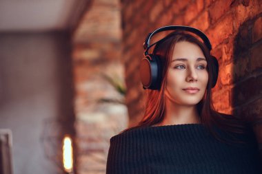 Yakın çekim portre Siyah kazaklı bir güzel şirin esmer çatı iç ile bir odada bir tuğla duvara yaslanmış iken iyi bir kulaklık ile müzik dinler.