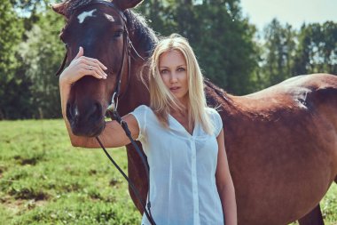 Beyaz bir bluz ve yeşil bir alana bir at sarılma kot giyen sevimli güzel sarışın.