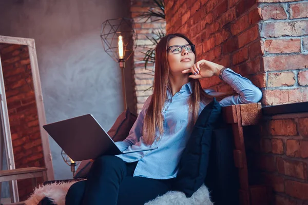 眼鏡と青シャツのラップトップでオンライン作業し ロフト デザイン ルームでソファの上に座っての魅力的なブルネット女性実業家の肖像画 — ストック写真