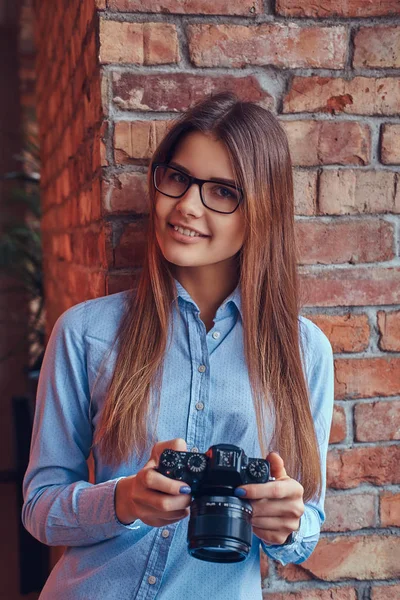 一个年轻的女摄影师在眼镜和蓝色衬衫上微笑着靠在一个房间里的砖墙上的阁楼设计的肖像 — 图库照片