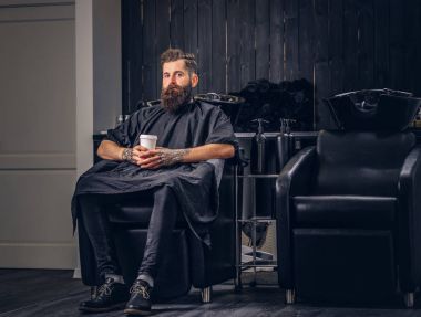 Saç yıkama önce bir kuaför salonu yakışıklı sakallı adam kollarını bir dövme ile kahve içer.
