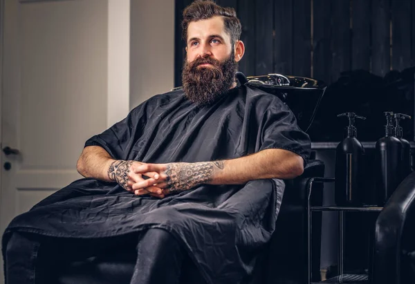 Handsome bearded male with a tattoo on his arms before hair wash in a hairdressers salon.