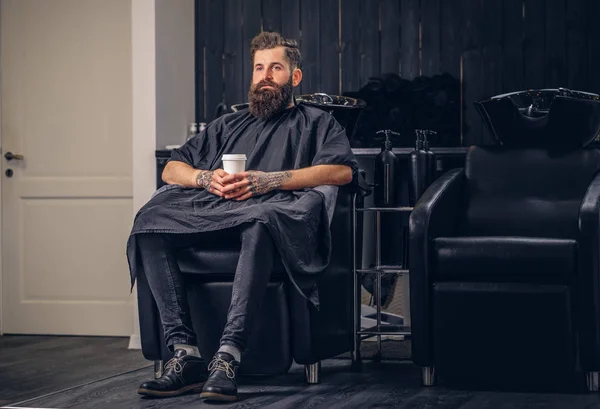 Handsome bearded man with a tattoo on his arms drinks coffee before hair wash in a hairdressers salon.
