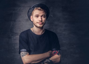 Çapraz dövme kollu yakışıklı sakallı hippi portresi bir siyah t-shirt ve şapka giymiş. Karanlık bir arka plan üzerinde izole.