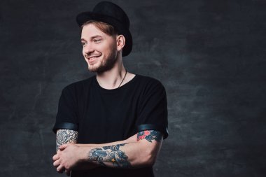 Çapraz dövme kollu yakışıklı sakallı hippi portresi bir siyah t-shirt ve şapka giymiş. Karanlık bir arka plan üzerinde izole.