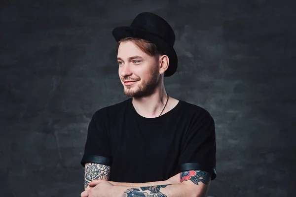 Portrait of handsome bearded hipster with crossed tattooed arms, dressed in a black t-shirt and hat. Isolated on a dark background.