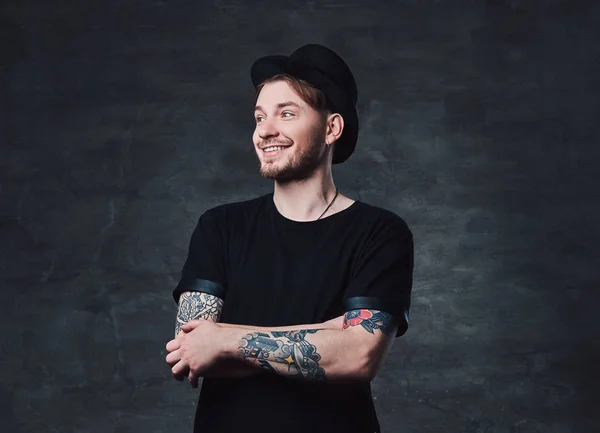 Portrait of handsome bearded hipster with crossed tattooed arms, dressed in a black t-shirt and hat. Isolated on a dark background.